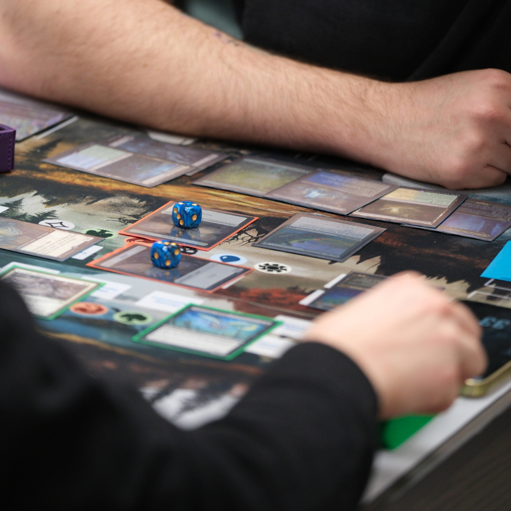 Zwei Personen spielen ein Sammelkartenspiel mit ausgelegten Karten und blauen Würfeln auf einer Spielmatte.